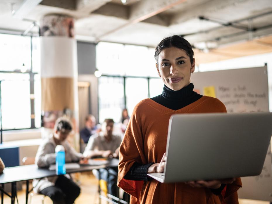 A teacher in her 20s holding a laptop smiling at the camera while her students work at desks behind her