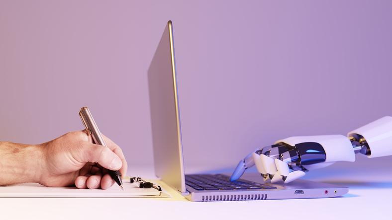 A human hand writes in a notebook while a robot hand types on a computer