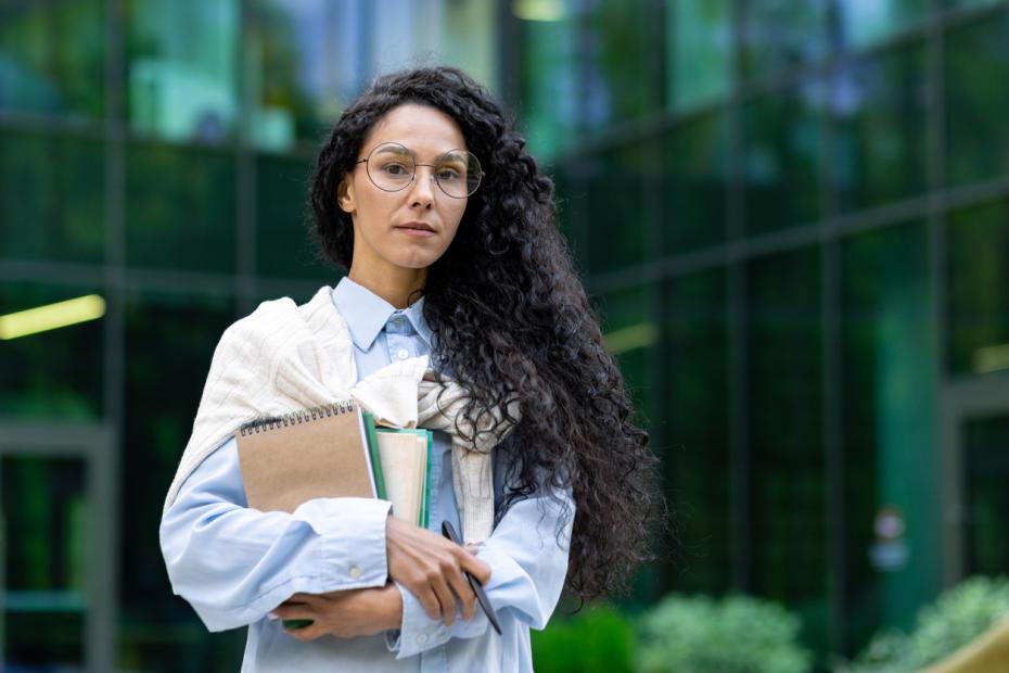 Female professor holding a pile of books standing in front of a university building