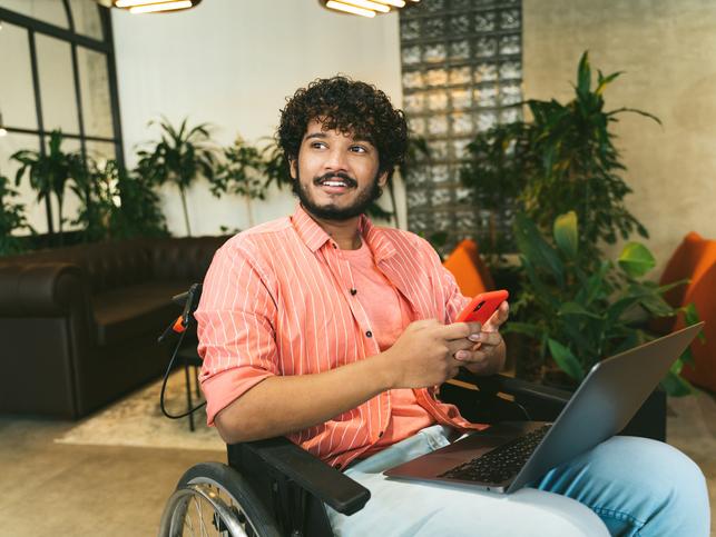 A young Asian man in a wheelchair uses a phone and a laptop