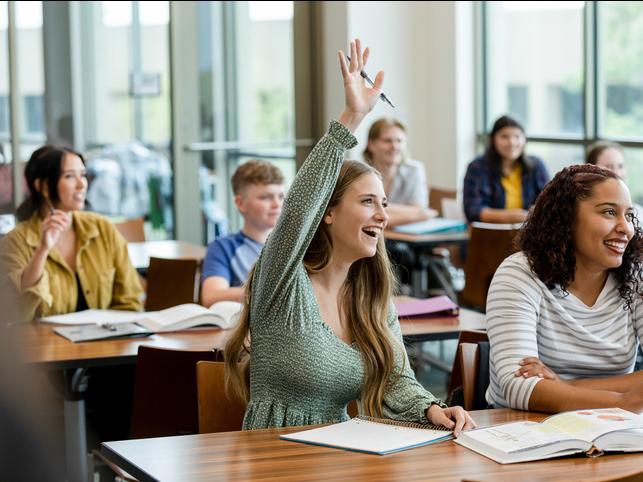 Students in class watching a teacher, among them one raises her hand to answer a question