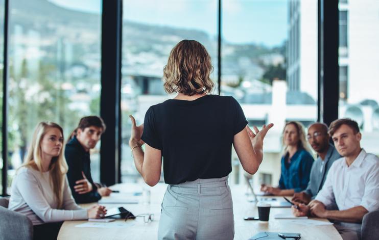 A woman stands in front of a boardroom table, delivering a speech to colleagues