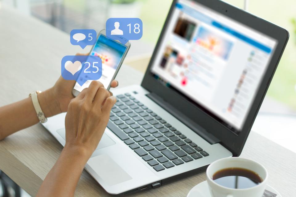 A laptop and a hand scrolling social media on a phone, surrounded by visualisations symbolising likes, comments and followers