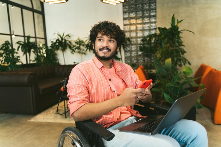 A young Asian man in a wheelchair uses a phone and a laptop