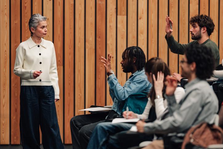 A professor explaining course structure to a class full of students, some of whom have their hands raised