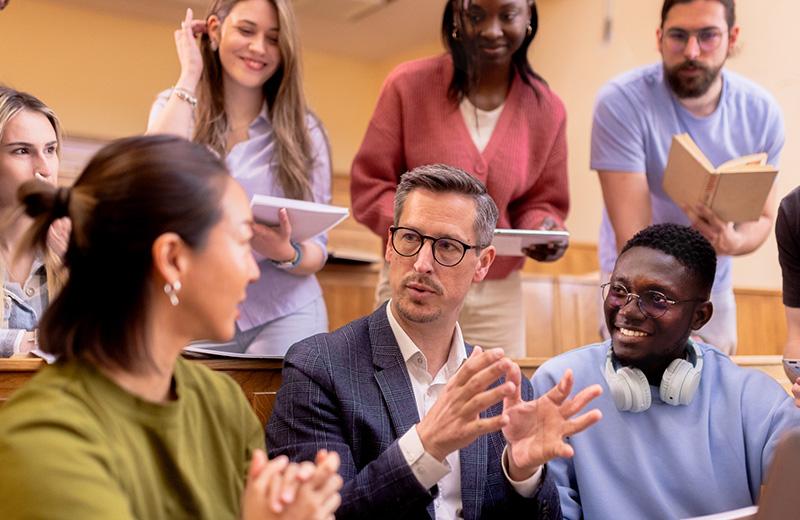 White male professor in discussion with diverse group of students in university amphitheatre