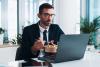 Businessman doing online study at laptop while eating takeaway lunch